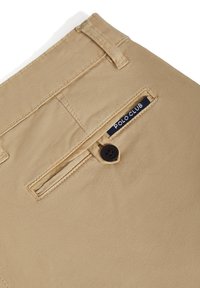 Beiga bomullsnickarbyxor med en ficklock bak med en svart knapp och en "POLO CLUB"-etikett i vit text på en marinblå bakgrund.