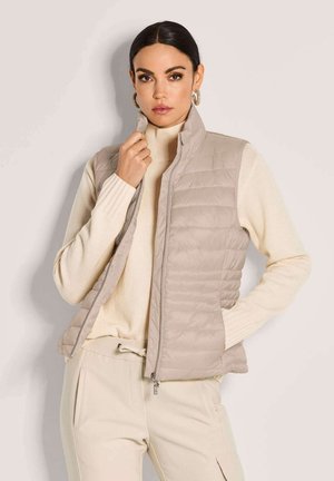 Frau mit dunklem Haar, die Creolen trägt, beige gesteppte Weste über einem cremefarbenen Rollkragenpullover und beige Hose mit den Händen in den Taschen.