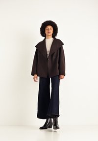 Cappotto oversize marrone con ampio colletto, abbinato a un maglione leggero a coste e jeans scuri a zampa d'elefante. Gli stivali neri con tacco completano l'outfit.