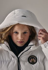 Giacca imbottita bianca con cappuccio grande e zip nera. Presenta una toppa rotonda con logo in tonalità di nero, rosa e oro sul petto.