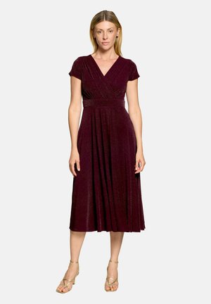 Femme portant une robe midi bordeaux scintillante à manches courtes, col en V et jupe plissée, debout en sandales neutres devant un fond uni.
