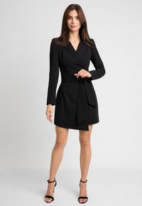 Czarna sukienka o kroju kopertowym z długimi rękawami, dekoltem w serek i paskiem w talii. Materiał wydaje się gładki, z krótką spódnicą i minimalistycznym designem.