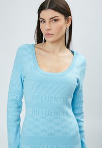 Lyseblå strikket sweater med en scoop-halsudskæring og lange ærmer, prydet med et struktureret "INFLUENCER" mønster gennem hele tøjet.