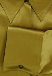 Koszulka z satyny w oliwkowym kolorze, z wykładanym kołnierzykiem, dużą kieszonką na piersi oraz zapięciem na guzik w kieszeni, z gładką, błyszczącą teksturą.