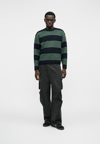 Maglione a righe in verde e blu navy con accenti scuri, abbinato a pantaloni cargo neri larghi. Il tessuto sembra morbido e testurizzato.