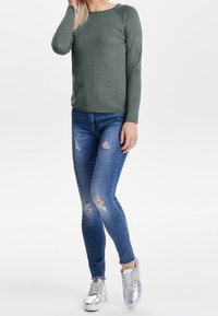 Grünes Langarmoberteil, schmal geschnittene blaue Ripped Jeans und silberne Sneaker, mit Rundhalsausschnitt und Raglanärmeln.