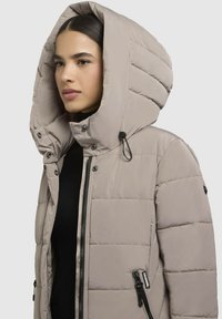 Žena nosí svetlo béžovú prešívanú bundu s veľkou kapucňou a čiernym zipsom cez čierne tričko, otočená doľava.