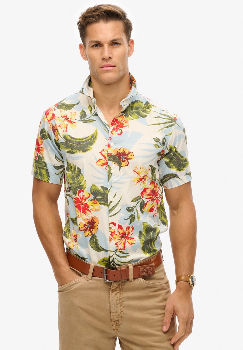 Camisa de manga corta con estampado floral que presenta un fondo azul claro, flores rojas y amarillas vibrantes, hojas verdes y un cuello con botones.
