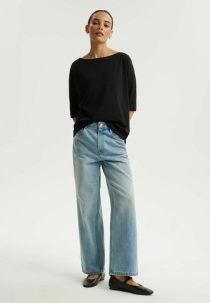 Kobieta stojąca, ubrana w luźną czarną bluzkę, jasnoniebieskie dzwoniaste jeansy i czarne baleriny na białym tle.