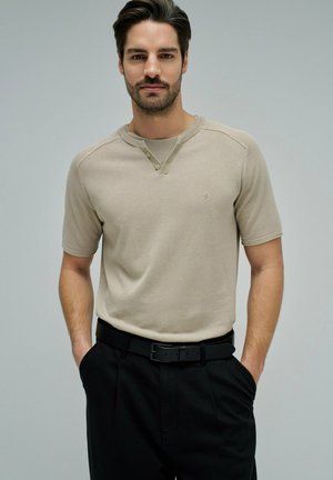 Homme barbu portant une chemise henley beige à manches courtes rentrée dans un pantalon noir avec ceinture noire, mains dans les poches, fond gris neutre.