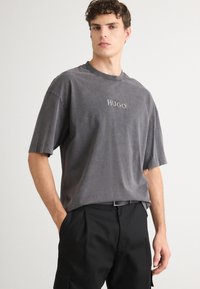 Graues Baumwoll-T-Shirt mit Rundhalsausschnitt und kurzen Ärmeln, mit einem gestickten "HUGO"-Logo auf der Vorderseite. Getragen mit schwarzen Hosen.