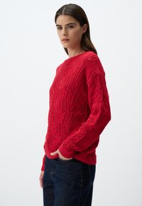 Maglione rosso lavorato a maglia con un motivo testurizzato e dettagli di cucitura aperta. Presenta un collo rotondo e maniche lunghe, leggermente sovradimensionate.