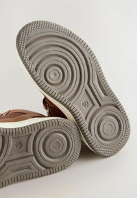 Brązowe skórzane podeszwy sneakersów z okrągłym wzorem bieżnika i rowkami. Szara guma z teksturowanym wykończeniem i zaokrąglonymi akcentami. Oznaczenie rozmiaru 12.