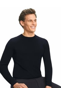 Schwarzer Rundhals-Pullover aus glattem Strickmaterial, mit langen Ärmeln und gerippten Bündchen, auf einem sitzenden Modell vor einem weißen Hintergrund präsentiert.