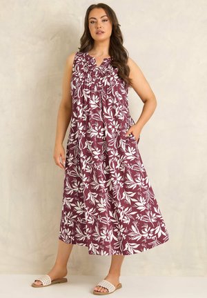Woman wearing sleeveless maroon midi dress with white floral print and white slip-on sandals, standing against a neutral wall.