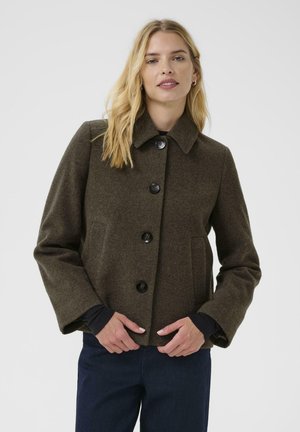 Femme blonde portant un manteau en laine olive foncé boutonné avec poches latérales et pantalon sombre sur un fond gris uni.
