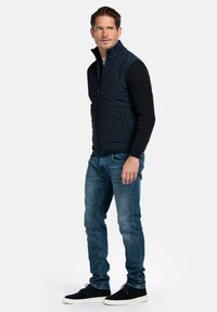 Marineblaue Steppweste über einem schwarzen Pullover, kombiniert mit schlanken blauen Jeans und schwarzen Sneakers. Verfügt über einen Reißverschluss und gerippte Details.