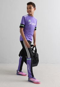 Zēns purpura un melnā futbola tērpā, turot melnu sporta somu, stāv pie vienkāršas gaiši pelēkas sienas.