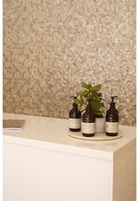 Three dark amber bottles with pump tops on a round marble tray, accompanied by a small green plant, on a glossy white surface. Textured gray wall.