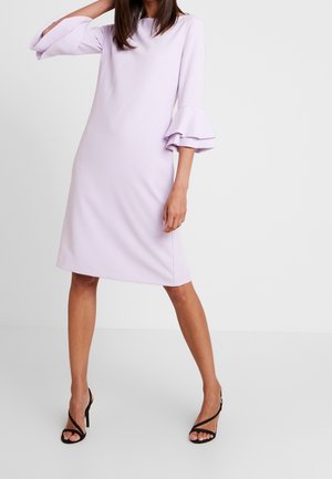 Mujer con un vestido lavanda hasta la rodilla, mangas acampanadas en capas y sandalias negras de tacón con tiras, posando delante de un fondo liso.