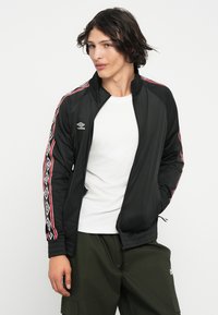 Črna zip-up jakna z rdečimi in belimi geometrijskimi vzorci ob rokavih, z visokim ovratnikom in elastičnimi manšetami, izdelana iz gladkega materiala.