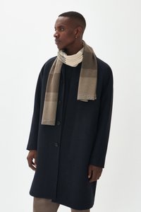 Homme portant un manteau boutonnière noir et une écharpe beige à motif, regardant à gauche sur un fond clair.