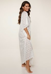 Cubre hombros estilo kimono de encaje blanco con mangas cortas, que presenta un patrón geométrico y un diseño suelto y fluido, que llega a la mitad de la pantorrilla.