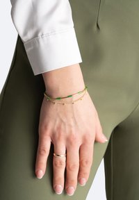 Złote i zielone bransoletki na nadgarstku, z płaskimi zielonymi koralikami i łańcuchowym wzorem z małymi elementami dekoracyjnymi. Widoczny mankiet gładkiej białej koszuli.