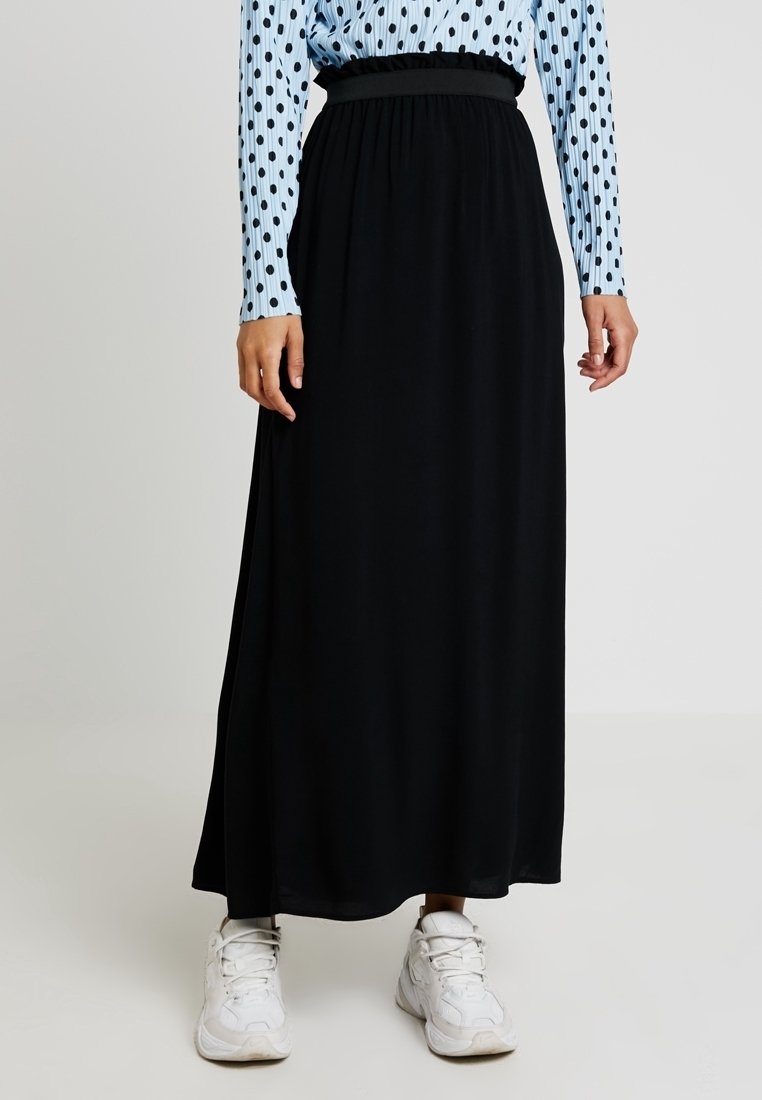 Femme portant une longue jupe noire avec une taille élastique, des baskets blanches et un haut à manches longues bleu clair à pois noirs.
