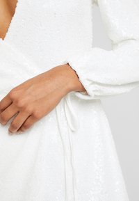 Main reposant sur la taille d'une robe blanche à sequins avec des manches longues et un détail noué à la taille.