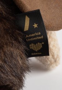 Brown plush fur material with a tan suede-like patch. Includes a black fabric label featuring gold text: "America Unlimited" and "CANADA Kollektion."