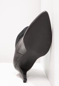 Chaussure à talon haut noire à bout pointu, inclinée contre un mur blanc sur une surface blanche, montrant la semelle et le talon fin.