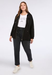 Donna che indossa una felpa nera con zip sopra una t-shirt bianca con grafica, jeans neri a vita alta e sneakers bianche, in piedi su uno sfondo bianco.