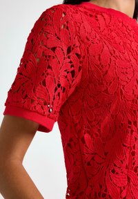 Kvinde iført en lys rød kortærmet bluse i blonder med intrikate blomster mønstre og en ensfarvet rød manchet på ærmet.