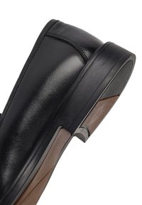 Schwarzer Lederschuh mit glänzender Oberfläche, ausgestattet mit einem niedrigen Absatz, strukturiertem Seitenpanel und kontrastierenden braunen Sohlenakzenten.