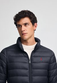 Granatowa kurtka puchowa z poziomymi przeszyciami, wysokim kołnierzem i czerwonym logo na lewym boku. Nosi ją h osoba w jasnym swetrze.