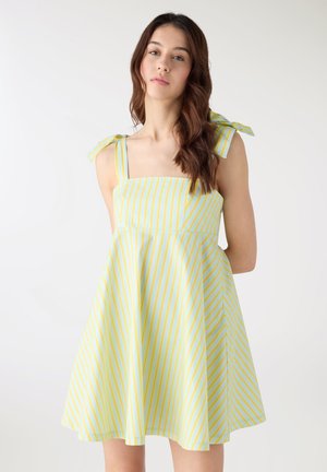 Donna con capelli castani lunghi che indossa un vestito senza maniche a righe azzurre e gialle con spalline annodate, in piedi contro uno sfondo semplice.