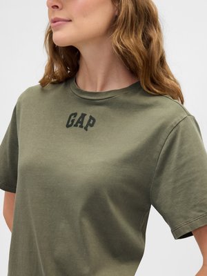 Kvinne i oliven grønn kortermet T-skjorte med "GAP" logo trykt i svart på brystet, som vender litt til venstre med synlige bølger i håret.