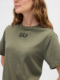 Olive zöld, rövid ujjú pólót viselő nő, amelyen fekete "GAP" felirat található a mellkasán, enyhén balra fordulva, látható hullámos hajjal.