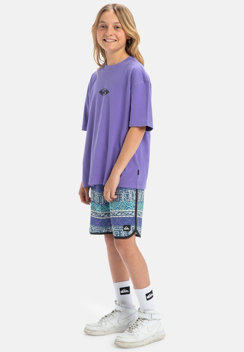 Camiseta oversized morada con un pequeño logo, pantalones cortos estampados en tonos azules y verdes, zapatillas blancas y calcetas blancas hasta la rodilla.
