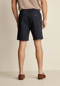 Námornické modré šortky s elastickým pasem, dvěma zadními kapsami na knoflíky a hladkým povrchem. Nosené s hnědými botami.