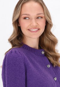 Violetti neulotty villatakki, jossa on korkea kaulus ja sydämen muotoiset hopeanväriset napit. Kankaan pinta on pehmeä ja tekstuuri on miellyttävä.