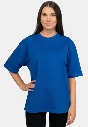 Jonge vrouw met lang bruin haar die een losse blauwe t-shirt draagt, staand met één hand op de heup tegen een effen lichte achtergrond.