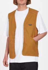 Homme portant une veste sans manches jaune moutarde à fermeture éclair sur un t-shirt blanc, avec une main dans la poche de la veste.