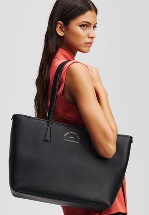 Borsa tote in pelle nera con finitura texturizzata, manici lunghi e logo inciso sul fronte. Modello mostrato con la borsa appoggiata sulla spalla.