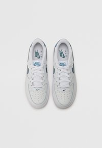Bijele Nike Air tenisice s glatkim kožnim gornjim dijelom, plavim detaljima, perforiranim prednjim dijelom i ravnim vezicama. Peta sadrži detalj s logotipom.