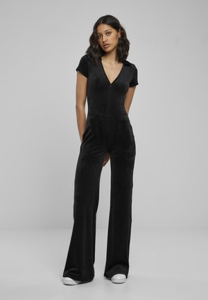 Kvinne står med hendene bak ryggen iført svart kortermet jumpsuit med frontglidelås og hvite joggesko mot en ensfarget bakgrunn.