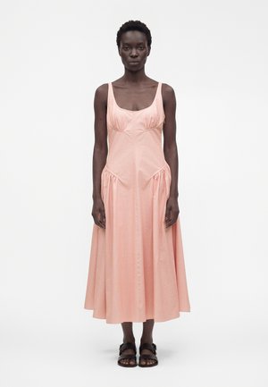 Kvinde med kort krøllet hår iført en ærmeløs, lys pink midi-kjole og sorte sandaler med spænder, stående mod en ensfarvet baggrund.