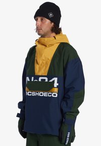 Windbreaker jacket featuring a yellow and navy design with a green accent. Notable front pocket and bold branding. Paired with black gloves.