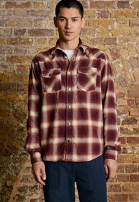 Young man wearing a red and beige plaid button-up shirt and navy pants standing against a textured brick wall background.